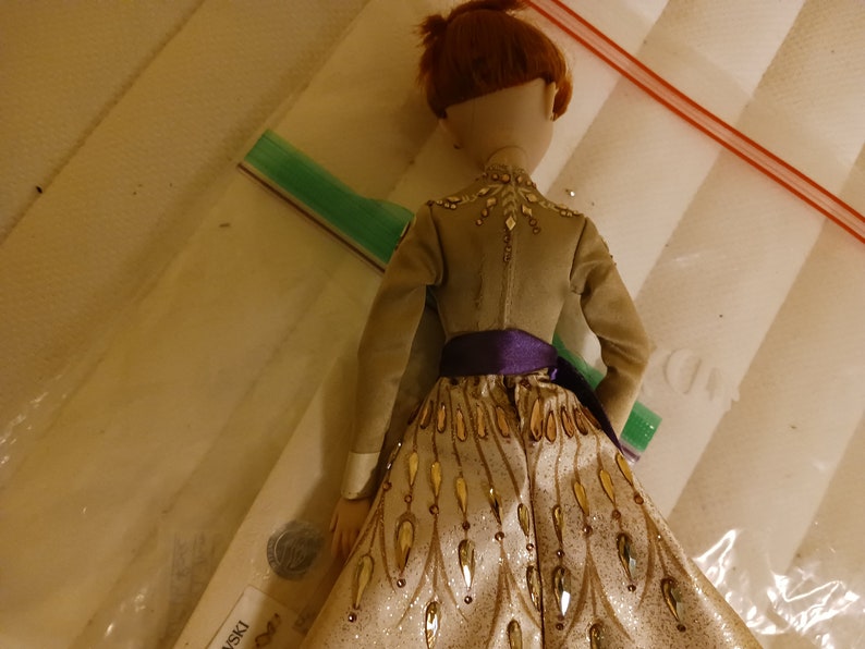May include: A doll wearing a gold and white dress with a purple sash. The dress has a sparkly, textured fabric and gold leaf accents. The doll has red hair and is wearing a brown jacket.