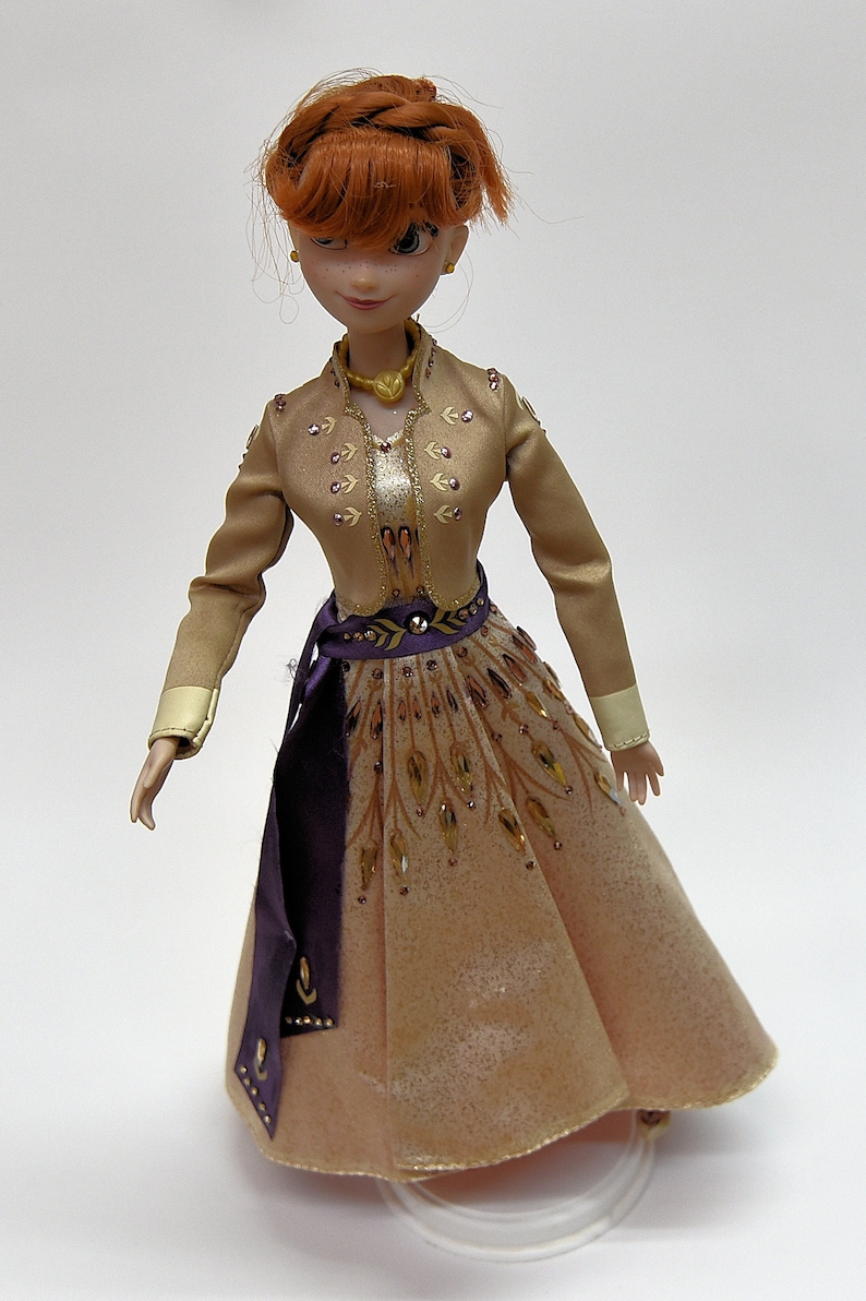 May include: A doll with red hair wearing a gold and brown dress with purple accents. The dress has a gold belt and intricate details.