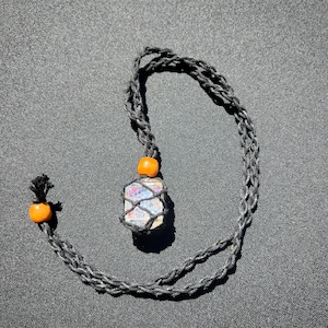 May include: A handmade necklace featuring a multi-colored crystal pendant encased in a black woven net. The necklace has a black braided cord with two orange wooden beads. The pendant is a mix of white, blue, and pink.