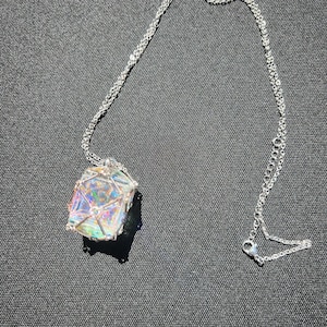 May include: A silver necklace featuring a geometric pendant with iridescent, multicolored crystal. The crystal is encased in a silver wire cage, and the chain is a delicate, linked design. The necklace is displayed on a dark gray surface.