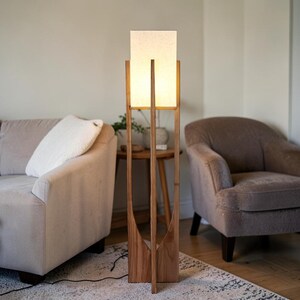 Puede incluir: Una l&aacute;mpara de pie de madera con una pantalla rectangular de color crema. La l&aacute;mpara tiene un acabado de madera natural y un dise&ntilde;o &uacute;nico de marco abierto. La l&aacute;mpara est&aacute; sobre una alfombra estampada en un sal&oacute;n.