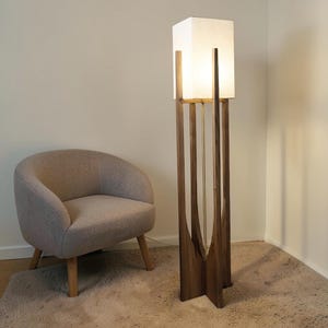 Puede incluir: Una l&aacute;mpara de pie moderna con una pantalla rectangular blanca y una estructura de madera oscura. La l&aacute;mpara tiene un dise&ntilde;o &uacute;nico con soportes verticales de madera y un acento de lat&oacute;n. Un sill&oacute;n beige est&aacute; cerca.