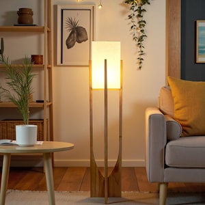 Puede incluir: Una l&aacute;mpara de pie de madera con una pantalla rectangular de color crema. La l&aacute;mpara tiene un dise&ntilde;o minimalista con tres soportes verticales de madera. La l&aacute;mpara est&aacute; en un sal&oacute;n con un sof&aacute;, una mesa auxiliar y plantas decorativas.