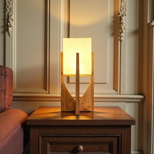 Puede incluir: Una l&aacute;mpara de mesa de madera con una pantalla cuadrada de color crema. La l&aacute;mpara tiene una base de madera geom&eacute;trica y un soporte vertical. Est&aacute; sobre una mesita de noche de madera oscura. La l&aacute;mpara est&aacute; encendida, emitiendo una luz c&aacute;lida.