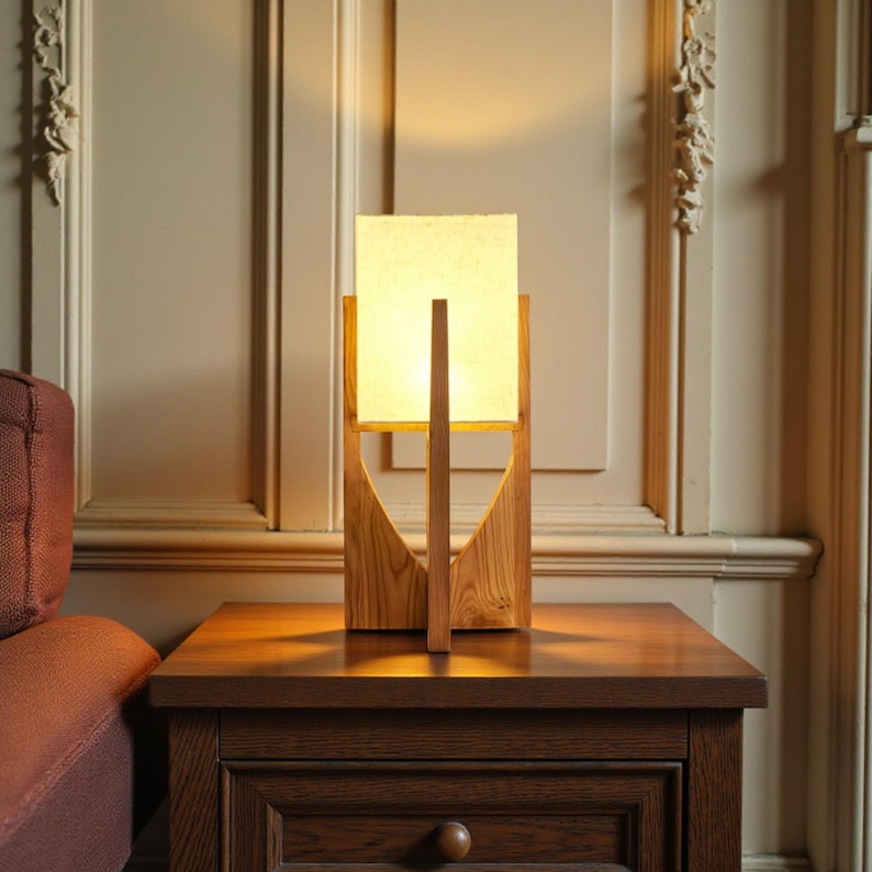 Puede incluir: Una l&aacute;mpara de mesa de madera con una pantalla cuadrada de color crema. La l&aacute;mpara tiene una base de madera geom&eacute;trica y un soporte vertical. Est&aacute; sobre una mesita de noche de madera oscura. La l&aacute;mpara est&aacute; encendida, emitiendo una luz c&aacute;lida.