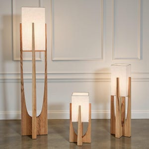 Puede incluir: Tres l&aacute;mparas de madera con pantallas rectangulares de color blanco roto. Las l&aacute;mparas tienen un acabado de madera natural y un dise&ntilde;o &uacute;nico de marco abierto. Las l&aacute;mparas var&iacute;an en altura, destacando la m&aacute;s alta. Las pantallas emiten un brillo suave y c&aacute;lido.