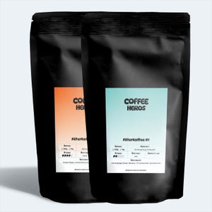 May include: Two black bags of coffee beans with white labels. The labels have the text "COFFEE HEROS" and information about the coffee, including the roast, origin, and taste notes. The bags are labeled "Filterkaffee" and "Filterkaffee #1".