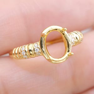 以下が含まれることがあります： 楕円形の中心部と小さな輝く石で飾られたバンドを備えた、金色のリングセッティング。 このリングは宝石を保持するように設計されています。 クローズアップビュー。