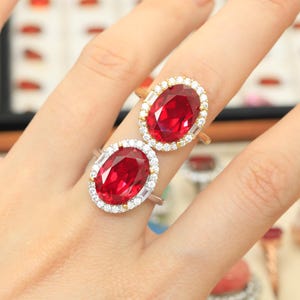 May include: Two rings featuring large oval-cut red gemstones as centerpieces. Each stone is surrounded by a halo of small, clear, round-cut stones and accented with baguette-cut stones on the sides, set in a metallic band.