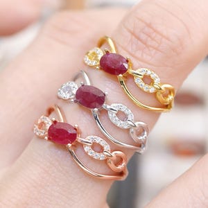 May include: Three rings with oval-cut ruby gemstones. The rings are in gold, silver, and rose gold tones. Each ring features a chain-link design with small, sparkling accents. The rings are displayed on a finger.