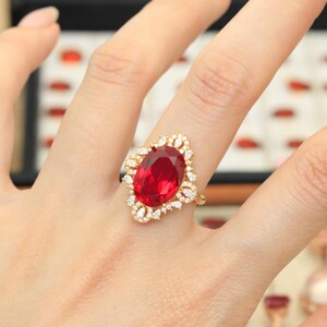 Puede incluir: Un anillo de oro con una gran piedra preciosa de color rojo intenso y corte ovalado como pieza central. La piedra central está rodeada por un halo de pequeñas piedras claras de corte marquesa y redondo en un engaste dorado festoneado.