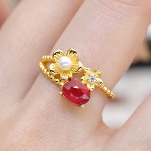 Op de afbeelding: Een gouden ring met een bloemmotief, een rode ovale edelsteen en een witte parel. De ring heeft ook een kleine diamant en een kralenband. De ring zit aan een vinger.