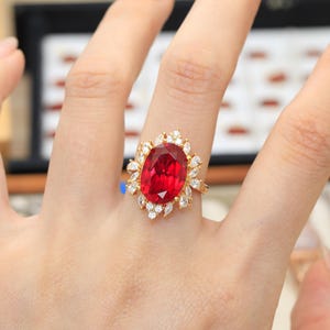 May include: A gold ring featuring a large, oval-cut red gemstone at the center, surrounded by a cluster of smaller, marquise-cut and round-cut clear white stones arranged in a sunburst pattern around the central red stone.