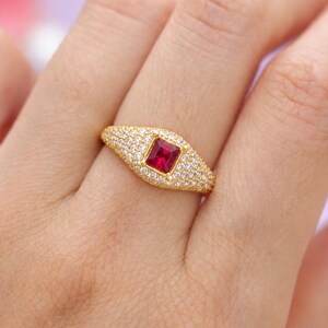 May include: A gold ring featuring a central rectangular red gemstone surrounded by a pavé setting of small clear stones. The band is wide and tapers slightly, with the entire surface of the ring covered in sparkling white stones around the ruby.