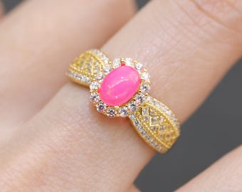 May include: A gold-toned ring featuring a vibrant pink oval gemstone at its center, surrounded by a halo of small, clear stones. The band has intricate detailing with additional clear stones, creating a luxurious and eye-catching design.