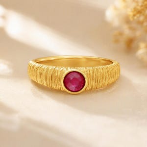 Puede incluir: Un anillo de oro con una banda texturizada de líneas verticales y una piedra preciosa de rubí redonda engastada en el centro. La superficie metálica tiene un acabado mate cepillado y la piedra de color rojo intenso está situada en el centro.