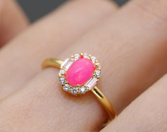 May include: A gold ring featuring a vibrant pink oval gemstone, surrounded by a halo of small, clear round and baguette-cut stones. The ring's band is a simple, polished gold.