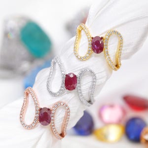 Puede incluir: Tres anillos con diseño de lazo, cada uno con un rubí ovalado central. Un anillo es dorado, otro plateado y otro de oro rosa. Las bandas están incrustadas con pequeñas piedras preciosas transparentes. Los anillos se exhiben sobre una tela blanca.