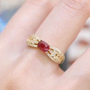 Op de afbeelding: Een gouden ring met een ovale robijn edelsteen in het midden. De ringband heeft een bladachtig filigraanontwerp, versierd met kleine, sprankelende diamanten. Dit is een sieraad.