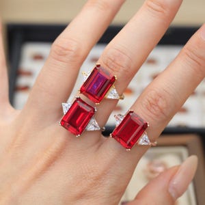 May include: Three rings featuring large, rectangular, emerald-cut red stones as the centerpieces. Each ring is accented by two smaller, triangular-cut clear stones on the sides. The bands are crafted from metallic materials in gold and silver tones.