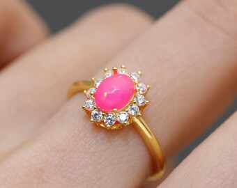 May include: A gold ring featuring a vibrant pink oval gemstone, surrounded by a halo of small, clear gemstones. The ring's band is a simple, polished gold. The setting is a classic, elegant design.