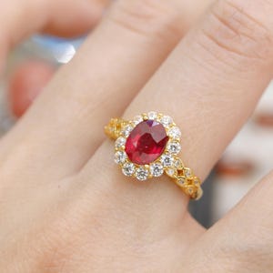 May include: A gold ring featuring a large, oval-cut ruby surrounded by a halo of small, round diamonds. The ring's band has a delicate, ornate design. The ruby is a deep red color, and the diamonds sparkle brightly.