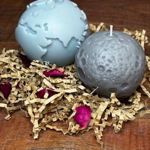 May include: Two round candles, one light blue and one gray, shaped like the Earth and the Moon. The candles are sitting on a bed of brown shredded paper with pink flower petals.