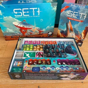 May include: A SETI board game set including a large box, a smaller Space Agencies expansion box, and an open game box containing various game components, including small colored tokens, card decks, and a game board with space-themed graphics.