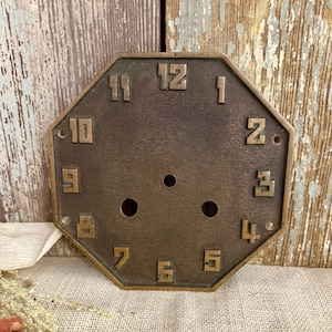 May include: An antique bronze clock face with an octagonal shape. The numbers 1 through 12 are embossed in a serif font. The clock face has three small holes near the center and is mounted on a wooden surface.