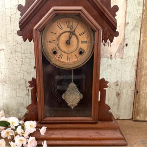 May include: Antique wooden clock with a pendulum and a key. The clock face has Roman numerals and a gold-colored finish. The clock is in a dark wood frame with intricate carvings.