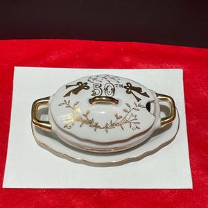 Puede incluir: Un recipiente de mantequilla de porcelana blanca con tapa y borde dorados. La tapa presenta un diseño dorado con el número 50 y la palabra "th".