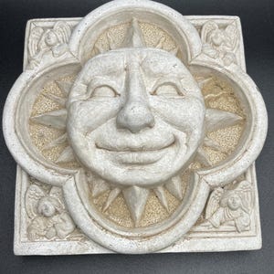 May include: A white ceramic wall plaque with a smiling sun face surrounded by four angels. The sun has a detailed face with a large nose and a wide smile. The angels are in a simple, stylized design.
