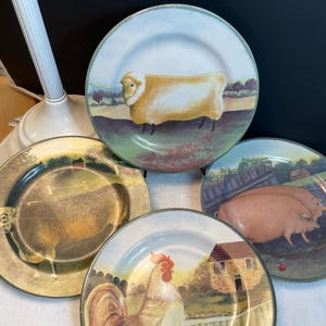 Puede incluir: Un juego de cuatro platos redondos de cerámica con ilustraciones de animales de granja. Un plato muestra una oveja blanca en un campo, otro dos cerdos rosas, un tercero un gallo cerca de una casa de piedra y el cuarto un cerdo en un pastizal.