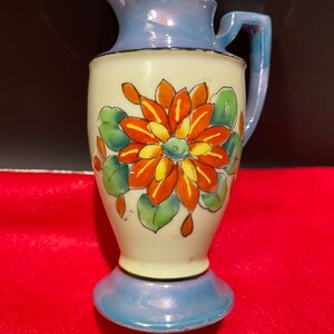 Op de afbeelding: Een kleine, blauwe en witte keramische kan met een bloemenmotief. De kan heeft een enkele oranje bloem met gele accenten en groene bladeren.