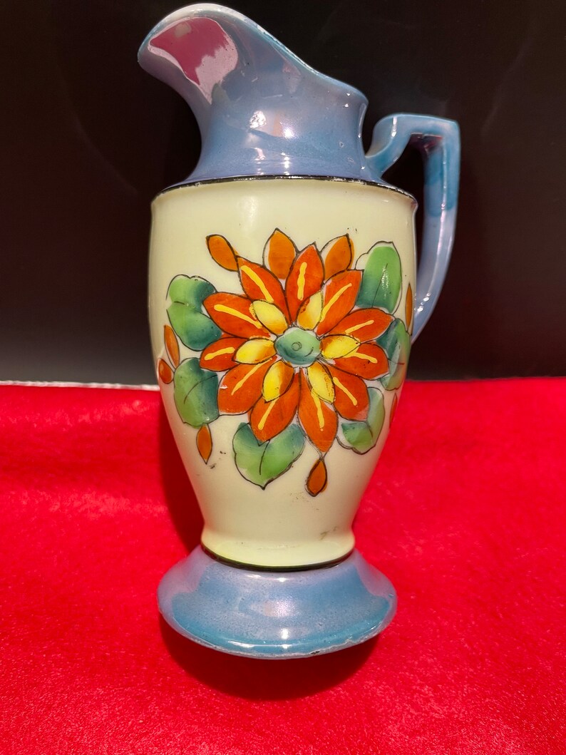 Op de afbeelding: Een kleine, blauwe en witte keramische kan met een bloemenmotief. De kan heeft een enkele oranje bloem met gele accenten en groene bladeren.