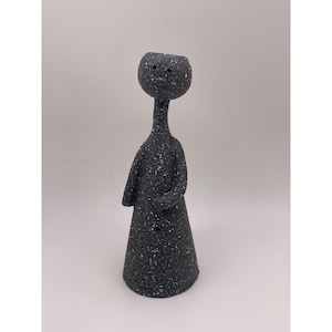 May include: A dark gray ceramic sculpture of a stylized figure with a round head and a long neck. The figure has a cone-shaped body and arms crossed in front. The surface is speckled with white dots.