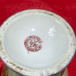 Op de afbeelding: Close-up van de onderkant van een witte keramische vaas met een rode en bruine stempel waarop "Hand Painted Made in Japan" en een gestileerd vogelontwerp staan.