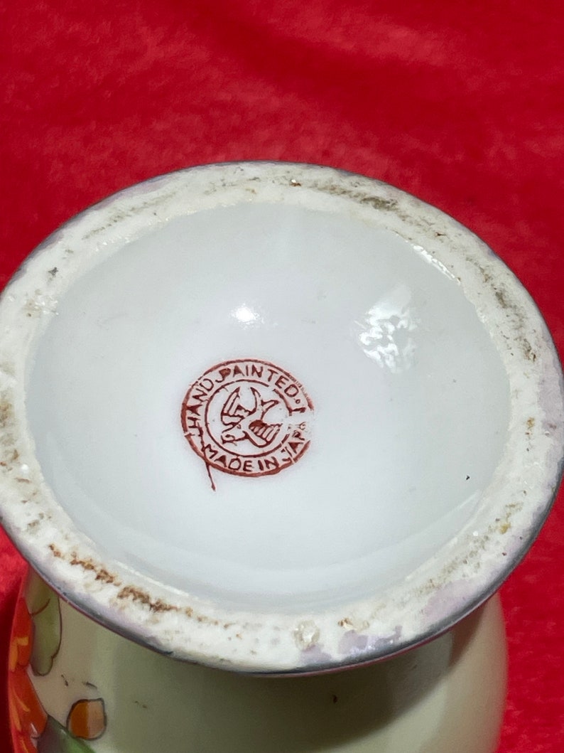 Op de afbeelding: Close-up van de onderkant van een witte keramische vaas met een rode en bruine stempel waarop "Hand Painted Made in Japan" en een gestileerd vogelontwerp staan.