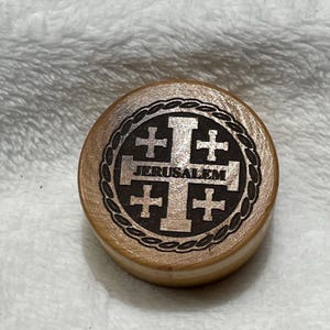 May include: A small wooden box with a round lid. The lid is decorated with a black and white image of a cross with four smaller crosses around it. The text "JERUSALEM" is printed below the cross.