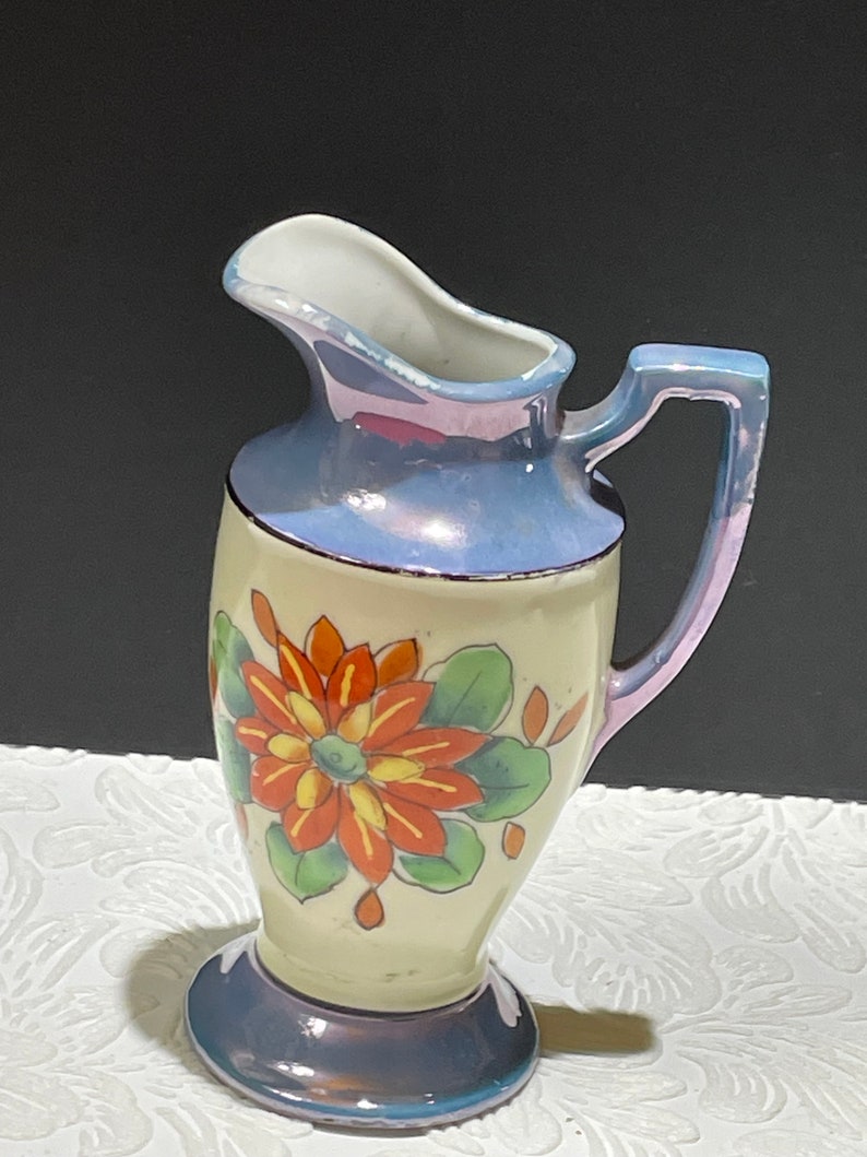 Op de afbeelding: Een kleine, witte keramische kan met een blauwe en iriserende glazuur. Het heeft een handgeschilderd bloemenmotief met oranje, gele en groene bloemen.