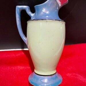 Op de afbeelding: Een kleine, vintage keramische kan met een lichtblauwe en witte glazuur. De kan heeft een rond lichaam en een uitlopende tuit. De handgreep is aan de zijkant van de kan bevestigd.