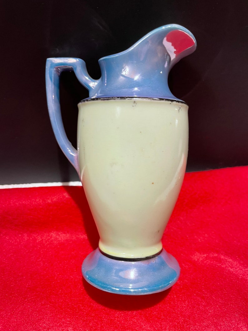 Op de afbeelding: Een kleine, vintage keramische kan met een lichtblauwe en witte glazuur. De kan heeft een rond lichaam en een uitlopende tuit. De handgreep is aan de zijkant van de kan bevestigd.