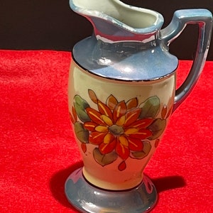 Op de afbeelding: Een kleine, blauwe en witte keramische kan met een bloemenmotief. De kan heeft een handvat en een brede opening. Het bloemenmotief heeft oranje en gele bloemen met groene bladeren.