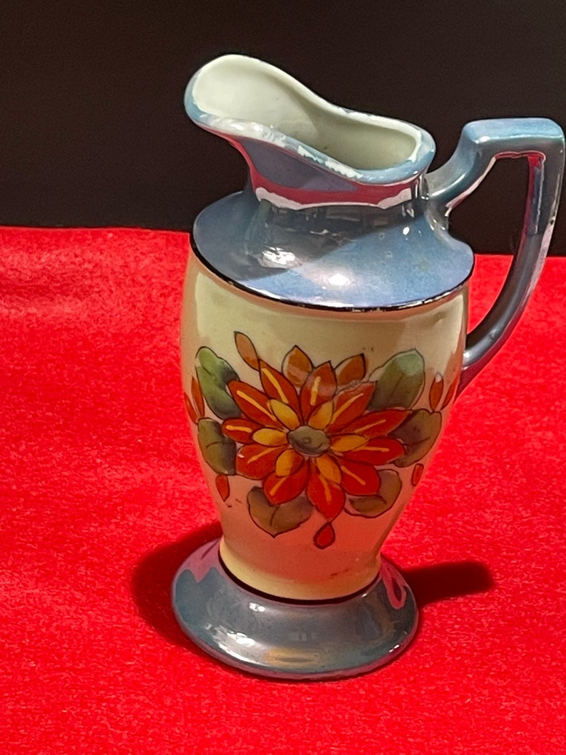 Op de afbeelding: Een kleine, blauwe en witte keramische kan met een bloemenmotief. De kan heeft een handvat en een brede opening. Het bloemenmotief heeft oranje en gele bloemen met groene bladeren.