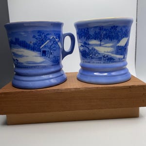 May include: Two blue ceramic mugs with a winter scene design. Each mug features a handle and a base. The mugs have a landscape illustration with a house and trees. Text on the mugs reads "Homestead in Winter" and "A Home in the Wilderness".