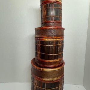 May include: A stack of three cylindrical dark brown bamboo storage containers with lids. Each container features vertical bamboo slats reinforced by horizontal bands and thin, light-colored binding cord, creating a tiered, rustic appearance.