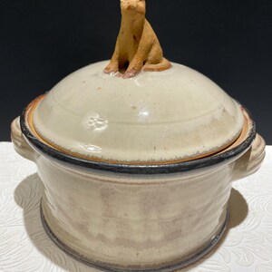 May include: A ceramic casserole dish with a lid featuring a small, brown cat figurine. The dish is off-white with a black rim and has two handles.