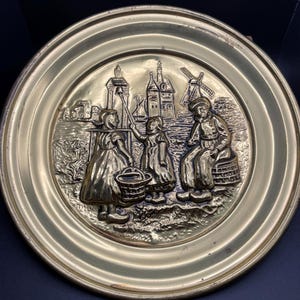 Puede incluir: Una placa de pared redonda de latón con un diseño en relieve de tres personas con ropa tradicional holandesa de pie frente a un molino de viento y edificios. La placa tiene un acabado dorado y un borde elevado.