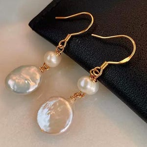 Può includere: Orecchini color oro con grandi perle bianche irregolari e perle rotonde più piccole. Gli orecchini hanno un design delicato avvolto con filo e una classica chiusura a gancio. Le perle hanno una lucentezza lucida.