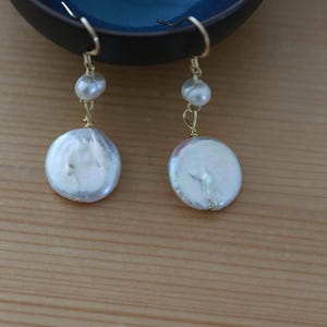 Könnte beinhalten: Elegante goldfarbene Ohrringe mit einem hängenden Design. Jeder Ohrring hat eine kleine, runde Perle oben, gefolgt von einer größeren, flachen, irisierenden Perle. Die Ohrringe sind vor einem verschwommenen Hintergrund platziert.